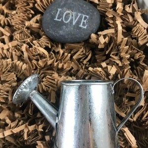 以下が含まれることがあります： 茶色の細断された紙の上に置かれた、ハンドルと注ぎ口が付いた小さな銀色のじょうろ。画像には、「LOVE」と刻まれた灰色の石も写っています。