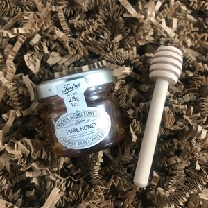 May include: A small glass jar of honey with a silver lid and label. The label reads "Tiptree" and "Pure Honey". The jar is sitting on a bed of brown paper shreds. A wooden honey dipper is next to the jar.