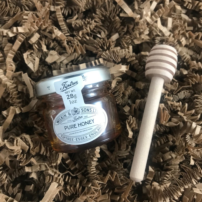 May include: A small glass jar of honey with a silver lid and label. The label reads "Tiptree" and "Pure Honey". The jar is sitting on a bed of brown paper shreds. A wooden honey dipper is next to the jar.
