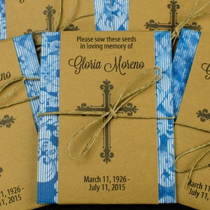 May include: Brown paper packets tied with twine and decorated with blue patterned paper. Each packet has a black cross and the text "Please sow these seeds in loving memory of Gloria Moreno" with the dates "March 11, 1926 - July 11, 2015".