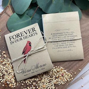 Peut inclure: Deux sachets de graines avec du texte et une illustration de cardinal, attach&eacute;s avec de la ficelle. L'un dit "Forever in Our Hearts" et comprend un nom et des dates. L'autre a un po&egrave;me sur un messager. Des graines sont &eacute;parpill&eacute;es.