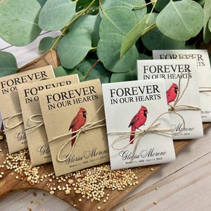 Peut inclure: Des sachets de graines avec une illustration de cardinal rouge et le texte "Forever in Our Hearts" sont attach&eacute;s avec de la ficelle. Ils sont pos&eacute;s sur une planche de bois avec des graines et des feuilles d'eucalyptus.
