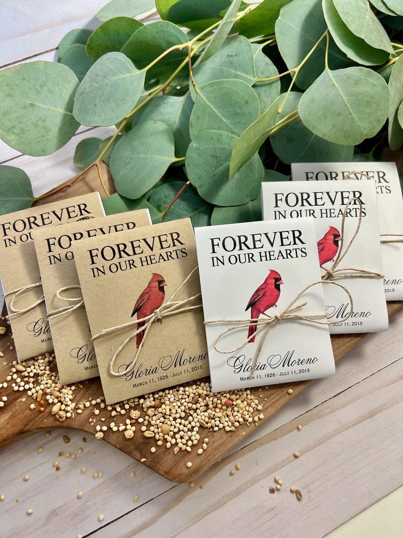 Peut inclure: Des sachets de graines avec une illustration de cardinal rouge et le texte "Forever in Our Hearts" sont attach&eacute;s avec de la ficelle. Ils sont pos&eacute;s sur une planche de bois avec des graines et des feuilles d'eucalyptus.