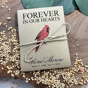 Peut inclure: Un sachet de graines beige avec le texte "FOREVER IN OUR HEARTS" et une illustration de cardinal rouge. Le sachet est nou&eacute; avec de la ficelle et porte le nom "Gloria Moreno" et les dates "11 mars 1926 - 11 juillet 2015".
