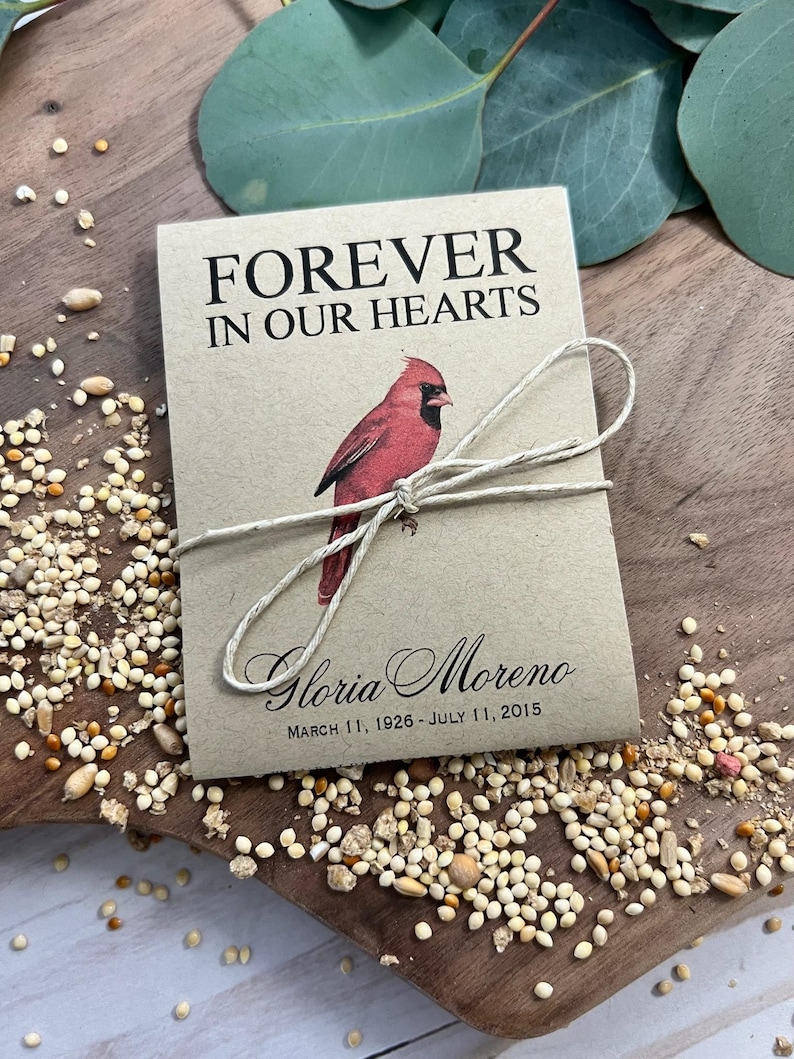Peut inclure: Un sachet de graines beige avec le texte "FOREVER IN OUR HEARTS" et une illustration de cardinal rouge. Le sachet est nou&eacute; avec de la ficelle et porte le nom "Gloria Moreno" et les dates "11 mars 1926 - 11 juillet 2015".