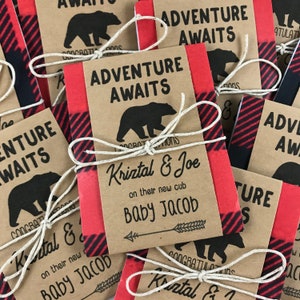May include: Red and black plaid favor boxes with a brown paper tag that says "Adventure Awaits" and "Kriztal & Joe on their new cub Baby Jacob".