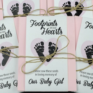 May include: Pink and white paper packets with the text "Footprints on our Hearts" and a pair of black footprints inside a pink heart. The packets are tied with twine and have the text "Please sow these seeds in loving memory of Our Baby Girl".