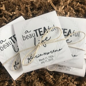 May include: Two white paper bags tied with twine. The bags have a black and white design with the text "a beautiful teaful life" and the name "Gloria Moreno" with dates.