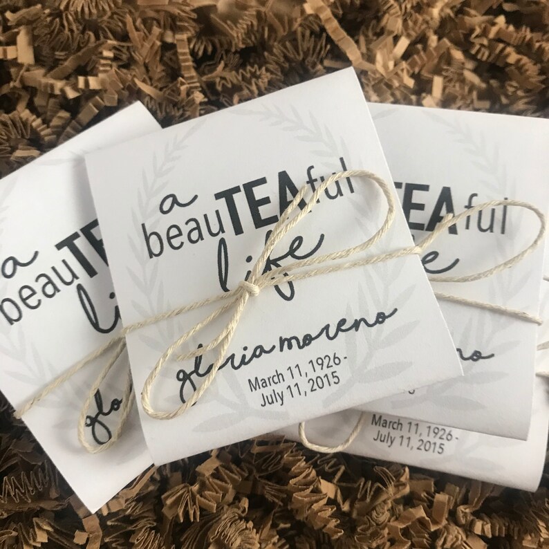 May include: Two white paper bags tied with twine. The bags have a black and white design with the text "a beautiful teaful life" and the name "Gloria Moreno" with dates.