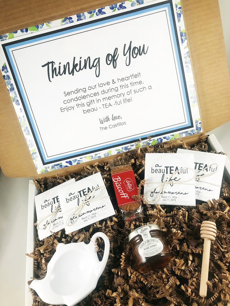 May include: A gift box filled with sympathy gifts for a loved one. The box contains a small white teapot, a jar of honey, a wooden honey dipper, a package of Biscoff cookies, and three tea bags with the text "a beautiful tea-ful life" and the name "Gloria Moreno".