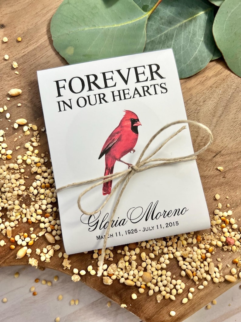 Peut inclure: Un paquet blanc avec le texte "FOREVER IN OUR HEARTS" et une illustration de cardinal rouge. Le paquet est nou&eacute; avec de la ficelle et comprend le nom "Gloria Moreno" et les dates "11 mars 1926 - 11 juillet 2015".