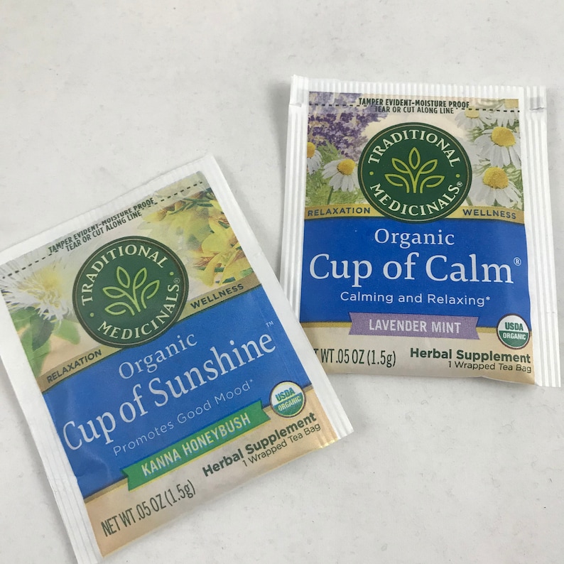 May include: Two individual tea bags, both labeled "Traditional Medicinals". The tea bag on the left is labeled "Organic Cup of Sunshine" with the text "Promotes Good Mood" and "Kanna Honeybush". The tea bag on the right is labeled "Organic Cup of Calm" with the text "Calming and Relaxing" and "Lavender Mint". Both tea bags are white with blue text and feature a green leaf logo.