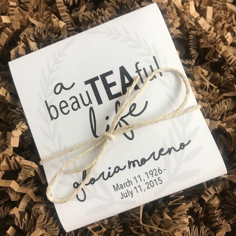 May include: A white card with a black wreath and the text "a beauTEAful life" and "Gloria Moreno" with the dates "March 11, 1926 - July 11, 2015". The card is tied with a natural twine bow.