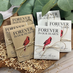 Peut inclure: Sachets de graines avec l'inscription "FOREVER IN OUR HEARTS" et une illustration de cardinal rouge. Chaque sachet est nou&eacute; avec de la ficelle et comprend le nom "Gloria Moreno" et les dates "11 mars 1926 - 11 juillet 2015".