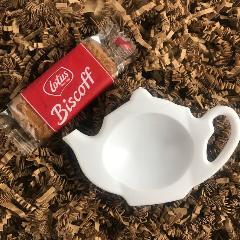 May include: A white ceramic teapot shaped like a traditional teapot, with a handle and spout. The teapot is sitting on a brown paper surface. A package of Lotus Biscoff cookies is also in the image.