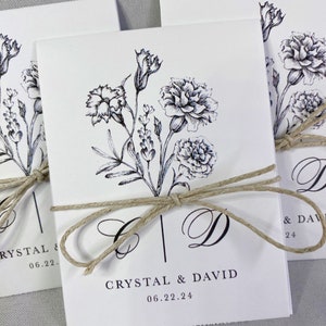 May include: White wedding invitation with black ink floral design and the text "Crystal & David 06.22.24" tied with a brown twine bow.