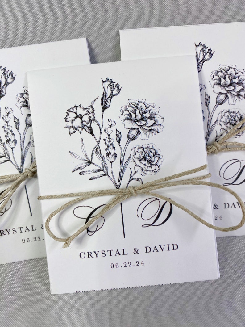 May include: White wedding invitation with black ink floral design and the text "Crystal & David 06.22.24" tied with a brown twine bow.