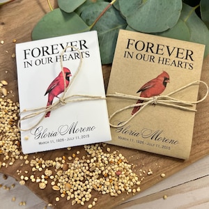 Peut inclure: Deux sachets de graines avec le texte "FOREVER IN OUR HEARTS" et une illustration de cardinal rouge. Les paquets sont attach&eacute;s avec de la ficelle et incluent le nom "Gloria Moreno" et les dates "11 mars 1926 - 11 juillet 2015".