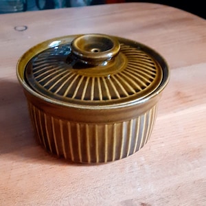 Puede incluir: Un plato de cerámica marrón con tapa. El plato tiene un diseño acanalado y una pequeña perilla en la tapa.