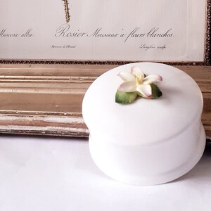 May include: A white porcelain trinket box with a delicate white flower with pink accents on the lid. The box is sitting on a white surface in front of a framed botanical illustration of a white rose with green leaves. The illustration is titled "Rosier Mousseux a fleurs blanches".