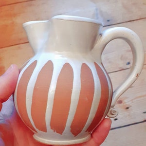 Op de afbeelding: Een witte keramische kan met een bruin en wit gestreept ontwerp. De kan heeft een handvat en een tuit.