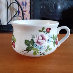 Op de afbeelding: Witte theekop van keramiek met een bloemenmotief met roze rozen, blauwe bloemen en groene bladeren. De kop heeft een ronde vorm en een handvat.