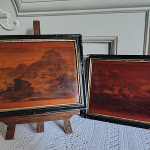 Könnte beinhalten: Zwei gerahmte Landschaftsbilder mit schwarzen Rahmen und goldenen Zierleisten. Ein Gemälde wird auf einer kleinen Holzstaffelei ausgestellt. Das Kunstwerk zeigt Berge und Gebäude in Orange- und Brauntönen.