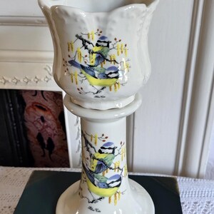 May include: A white ceramic candle holder with a scalloped rim, featuring a repeating illustration of two blue tits perched on branches with yellow catkins. The text "Blue Tit" is printed in small black script below each bird illustration.