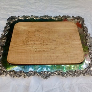 May include: A silver serving tray with an ornate, scalloped edge and a wooden cutting board insert. The tray has a silver finish and a decorative design.