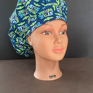 Könnte beinhalten: Eine blaue und grüne gemusterte OP-Mütze mit dem Wort "Star Wars", das sich über die gesamte Oberfläche wiederholt. Die Mütze zeigt eine kleine grüne Alienfigur.