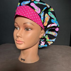 May include: A black scrub hat with a pink polka dot trim and a colorful flip-flop print. The hat is on a mannequin head.