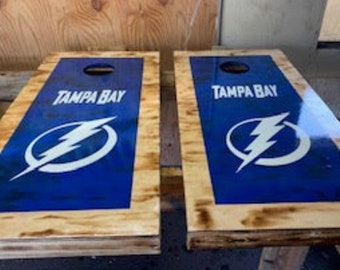 May include: Two wooden cornhole boards with a blue background and the Tampa Bay Lightning logo in white. The boards are made of wood with a natural finish and have a clear coat finish.