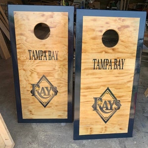 Puede incluir: Dos tableros de cornhole de madera con un acabado de veta de madera clara. Cada tablero tiene un borde negro, un agujero centrado y las palabras "TAMPA BAY" encima de un logotipo de "RAYS".