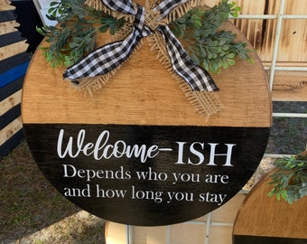 May include: A round wooden sign with a black and white checkered ribbon and greenery. The sign says "Welcome-ISH Depends who you are and how long you stay".