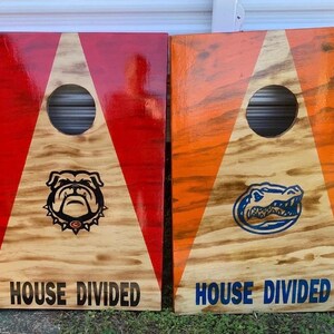 Könnte beinhalten: Zwei Cornhole-Bretter aus Holz. Eines ist rot mit einem Bulldoggen-Logo, das andere orange mit einem Alligator-Logo. Jedes Brett hat ein zentrales Loch und den Text "HOUSE DIVIDED" unten.