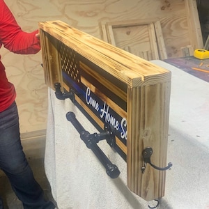 May include: Wooden wall-mounted shelf with a distressed American flag design, featuring a blue line. The shelf includes black metal hooks and the text "Come Home Safe".