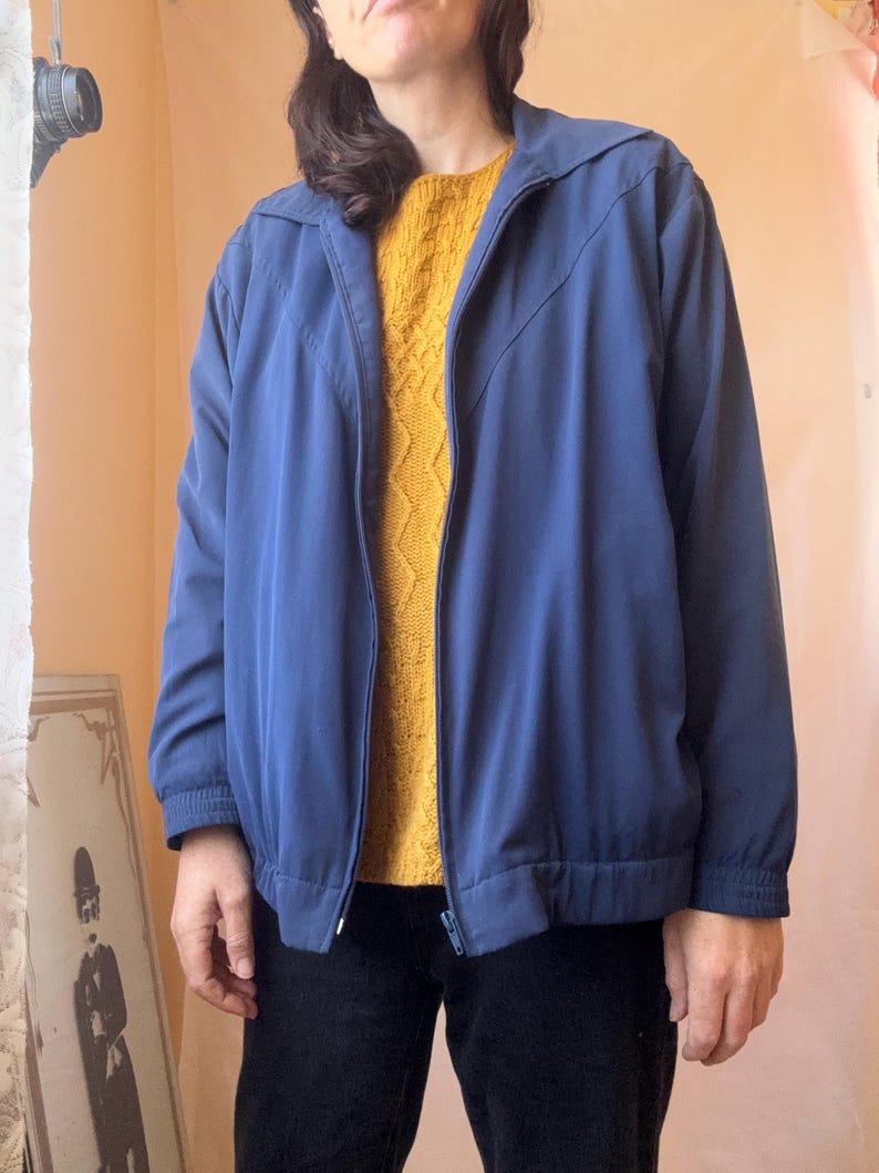May include: A solid blue zip-up jacket featuring a pointed collar, long sleeves with elastic cuffs, and a matching elastic waistband. The jacket has a front zipper closure and a distinct V-shaped seam detail across the chest area.