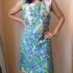 May include: A sleeveless knee-length dress featuring a white base with a vibrant floral pattern in shades of blue and green. The garment has a round neckline and a straight silhouette, showcasing a dense print of various flower shapes and leaves.