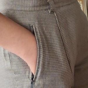 Puede incluir: Primer plano de un pantalón de pata de gallo gris y blanco con un bolsillo con cremallera. El pantalón tiene una cintura alta y un cierre de botón. Se ve una parte superior negra por encima del pantalón.