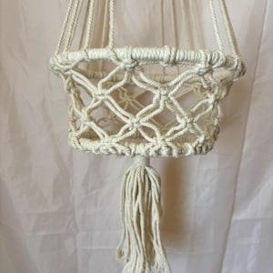 May include: An off-white macrame plant hanger constructed from thick cotton cord. The design features a circular knotted basket with a diamond-shaped lattice pattern, multiple suspension cords extending upwards, and a single decorative tassel at the base.