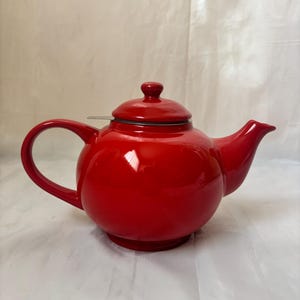 May include: A glossy, bright red ceramic teapot with a rounded body, a curved handle, and a matching lid. A small metallic tea infuser rim is visible beneath the lid. The teapot features a short, tapered spout and a stable, circular base.