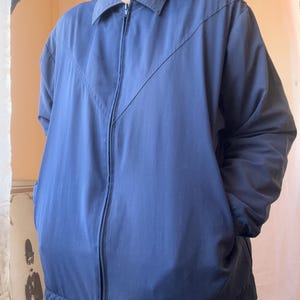 May include: A solid navy blue zip-up jacket featuring a classic collar, long sleeves, and a distinct V-shaped seam detail across the chest. The garment includes a full-length front zipper closure and two side pockets, providing a clean and functional design.