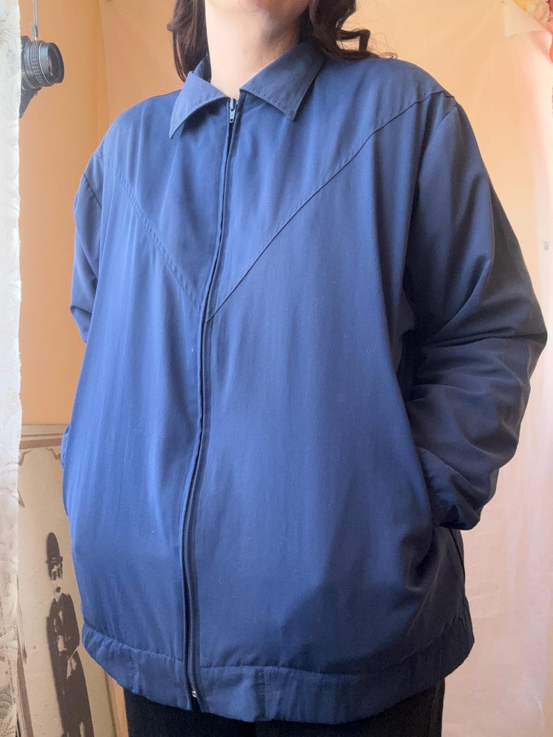May include: A solid navy blue zip-up jacket featuring a classic collar, long sleeves, and a distinct V-shaped seam detail across the chest. The garment includes a full-length front zipper closure and two side pockets, providing a clean and functional design.