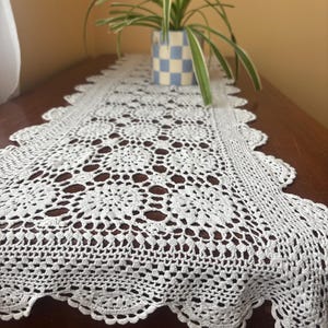 May include: A white, open-weave crochet table runner with a scalloped edge, displayed on a dark brown wooden surface. A small potted plant with green and white striped leaves is in the background.