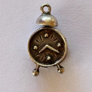 May include: A small silver-toned metal charm shaped like an alarm clock. The charm features a circular face with raised dot hour markers, a central V-shaped hand, two small feet at the base, and a bell-shaped top with a circular jump ring loop.