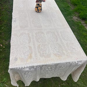Puede incluir: Un mantel rectangular de ganchillo color blanco roto con un patrón floral cuadrado repetitivo y detalles calados intrincados. Un pequeño jarrón de cerámica con estampado de flores negras y amarillas que contiene una planta descansa sobre la tela.