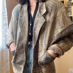 Op de afbeelding: Een bruin en beige leren blazer met een textuurpatroon. De blazer heeft een klassieke revers, twee voorzakken en een relaxte pasvorm. De mouwen zijn iets opgerold, waardoor een donkerdere bruine voering zichtbaar is. Gedragen met een zwart overhemd en een blauwe jeans.