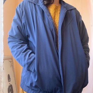 May include: A solid navy blue zip-up jacket featuring a pointed collar, long sleeves with ribbed cuffs, and a V-shaped seam detail across the chest. The jacket includes a full-length front zipper closure and an elasticized hem at the waistline.