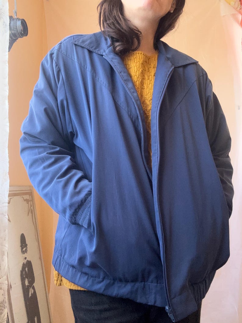 May include: A solid navy blue zip-up jacket featuring a pointed collar, long sleeves with ribbed cuffs, and a V-shaped seam detail across the chest. The jacket includes a full-length front zipper closure and an elasticized hem at the waistline.
