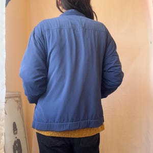 May include: A solid blue long-sleeved jacket with a collared neckline and a straight hemline. The jacket is worn over a mustard yellow textured sweater and dark-colored pants. The back view shows a horizontal seam across the upper back area.